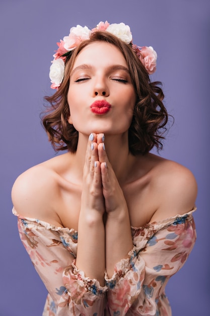 Веселая девушка в романтическом наряде позирует с закрытыми глазами. Фотография в помещении приятной кудрявой дамы в венке из цветов.