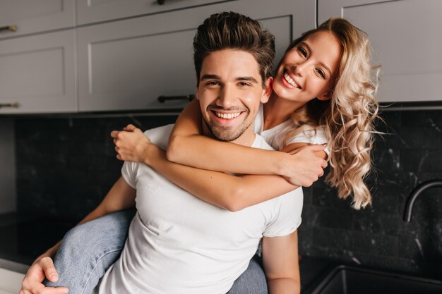 Jocund girl with curly hairstyle embracing husband. Smiling couple posing together in kitchen.