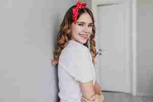 Free photo jocund stylish girl posing at home with smile. indoor photo of stunning female model wears red ribbon looking over shoulder.