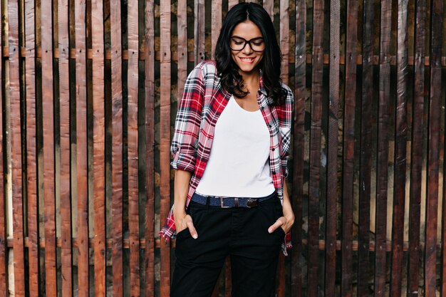 Jocund woman in black pants posing with hands in pockets. Outdoor photo of sensual latin female model wears casual attire.