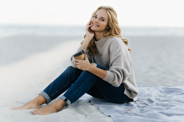 Jocund woman in jeans sitting at beach with cup of coffee. Attractive blinde woman posing in sand in autumn day.