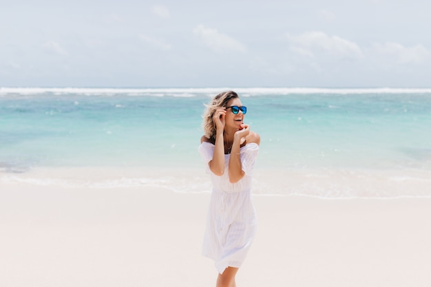 Free photo jocund woman in romantic white attire standing on sea. laughing ecstatic woman in sunglasses spending summer day at ocean.