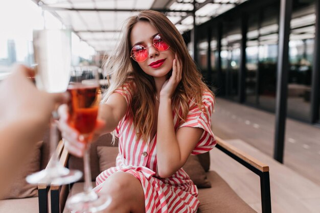 Jocund young lady in pink glasses relaxing in cafe with glass of cocktail Adorable blonde girl sitting in restaurant in good summer day