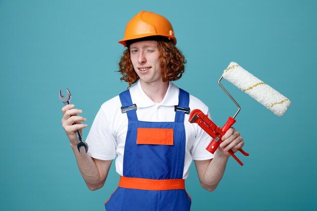 青い背景で隔離の制服を着たローラーブラシでレンチを保持しているJokin若いビルダー男