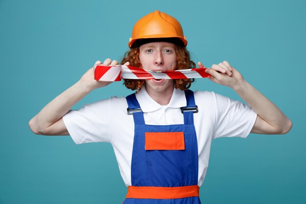 Jokin young builder man in uniform holding duct tape on mouth isolated on blue background