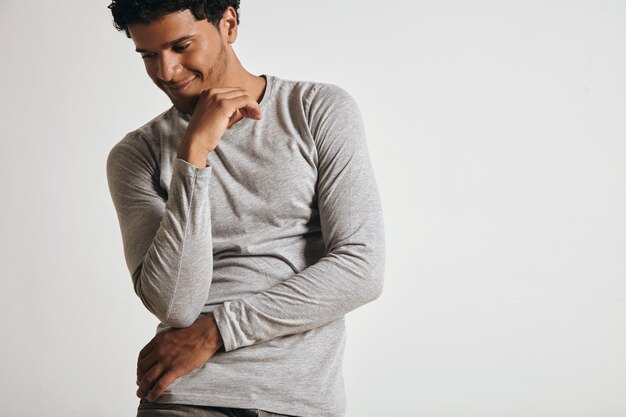 Jouyful smiling young latino guy in blank grey longsleeve looks at side and fposes in front of white wall