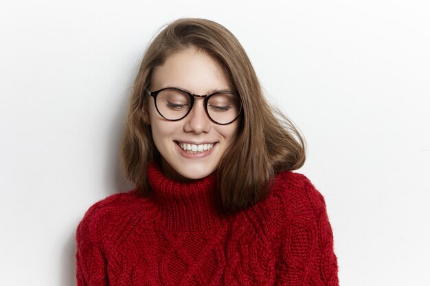 喜びと幸福の概念。透明な眼鏡とスタイリッシュな栗色のタートルネックのセーターを着て、恥ずかしがり屋の笑顔で見下ろし、恥ずかしがり屋の遊び心のある神秘的な若い白人女性の写真