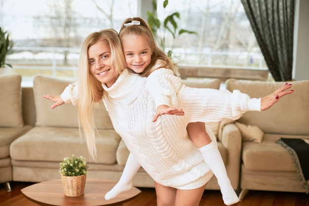 Joy, happiness and leisure time concept. Pretty girl with long hair having ride on her motherâs back, keeping arms outstretched. Young mom and daughter having fun in living room, fooling around