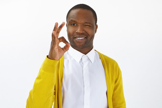 Joy, happiness and positiveness concept. Confident young dark skinned male employee expressing readiness to complete task, showing okay gesture and smiling cheerfully at camera, posing in studio wall