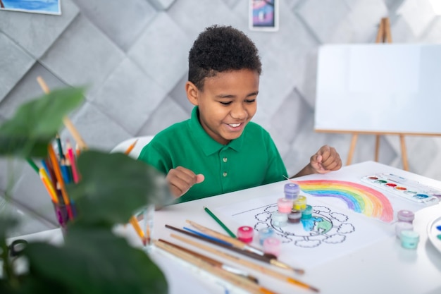 Free photo joy. happy dark-skinned boy of school age in casual clothes sitting at table joyfully looking at built pyramid of paints in light room with easel