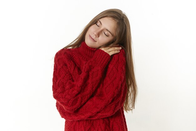 Концепция радости, релаксации и счастья. Красивая юная леди в уютном свитере с длинными рукавами, обнимая себя и закрывая глаза от удовольствия и удовольствия, счастливо улыбаясь, наслаждаясь теплой одеждой