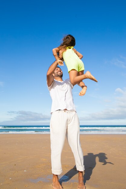 Joyful active dad holding little daughter in arms and lifting her up in air while spending time with girl on ocean beach
