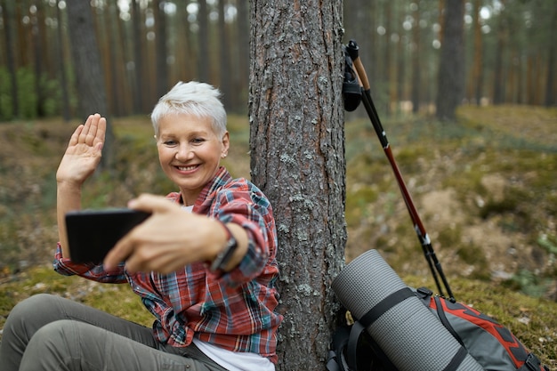 Free photo joyful active retired female sitting under tree with hiking gear holding cell phone, smiling and waving hand, talking to her friend via video conference call using online app.