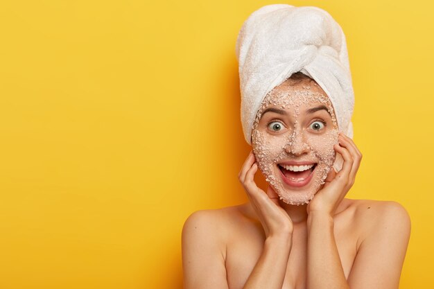 Joyful adorable woman with glad facial expression, applies natural sea salt mask, has healthy well cared skin, wears towel on head, enjoys hygienic treatments