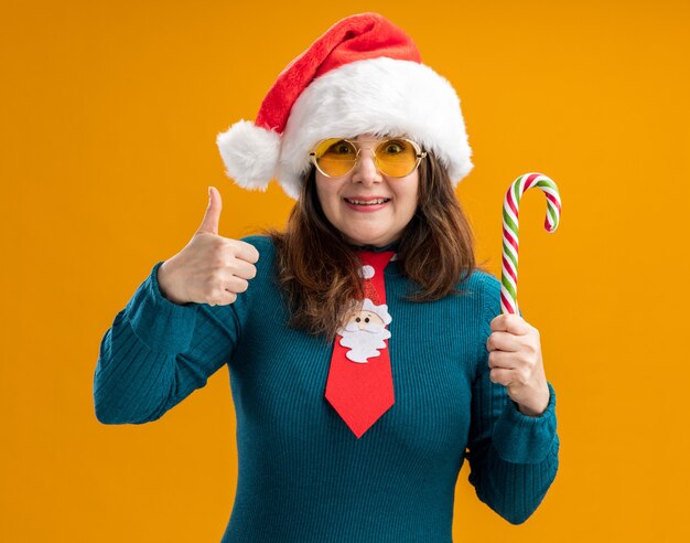 Joyful adult caucasian woman in sun glasses with santa hat and santa tie holds candy cane and thumbs up isolated on orange wall with copy space