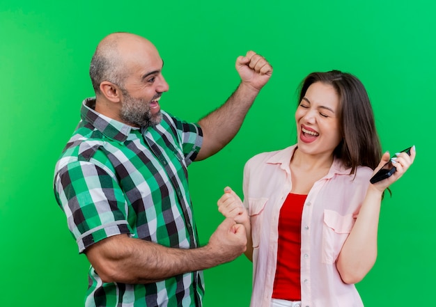 Joyful adult couple man standing in profile view woman holding mobile phone with closed eyes both clenching fists isolated on green wall