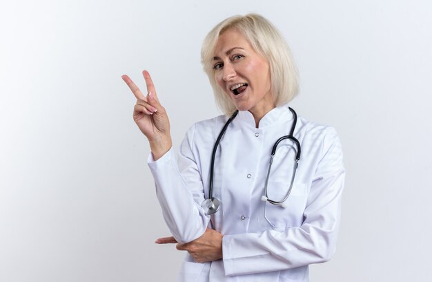 joyful adult female doctor in medical robe with stethoscope gesturing victory sign isolated on white wall with copy space