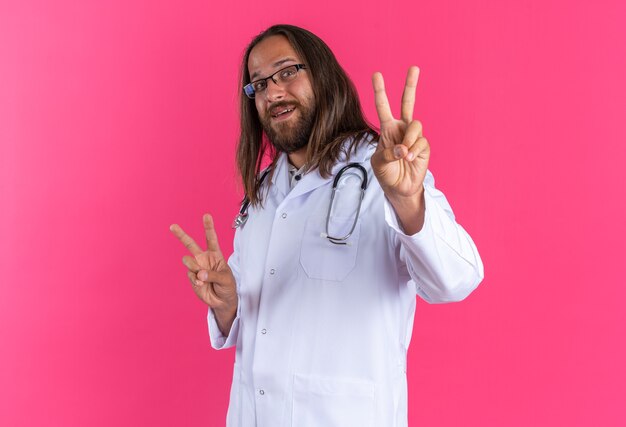 Joyful adult male doctor wearing medical robe and stethoscope with glasses standing in profile view doing peace sign looking at camera isolated on pink wall