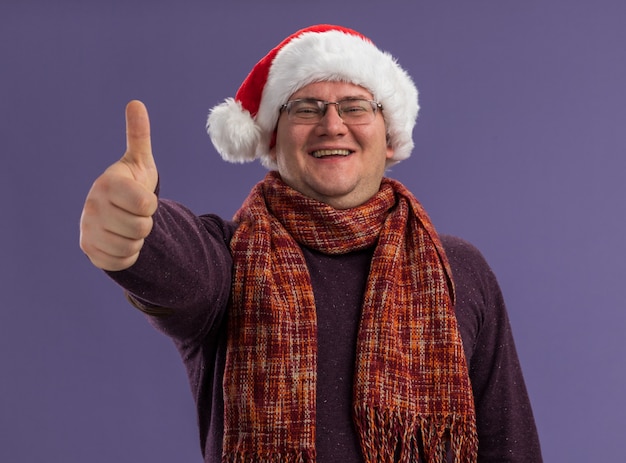 Foto gratuita gioioso uomo adulto con gli occhiali e il cappello di babbo natale con la sciarpa intorno al collo che mostra il pollice in alto isolato sul muro viola