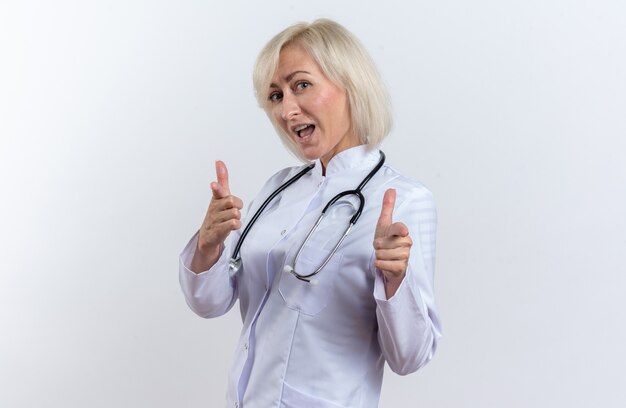 Joyful adult slavic female doctor in medical robe with stethoscope pointing at camera isolated on white background with copy space