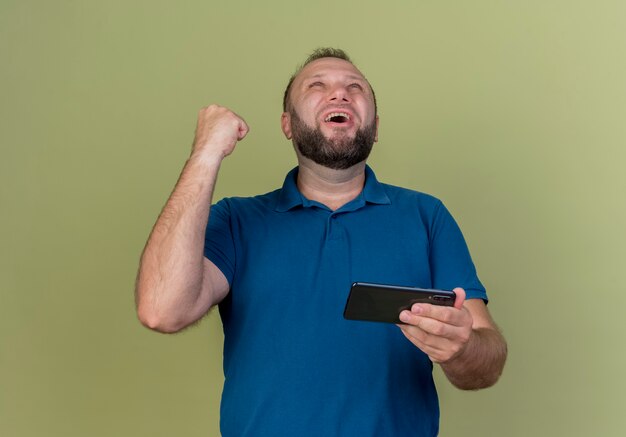Радостный взрослый славянский мужчина держит мобильный телефон, глядя вверх, делая жест да