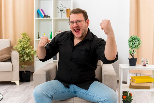 Joyful adult slavic man in optical glasses sits on armchair keeping fists up looking at camera inside the living room