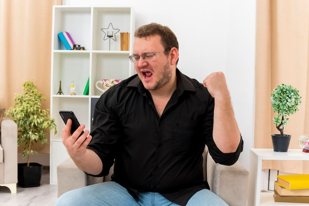 Joyful adult slavic man in optical glasses sits on armchair looking at phone and keeping fist up inside designed living room