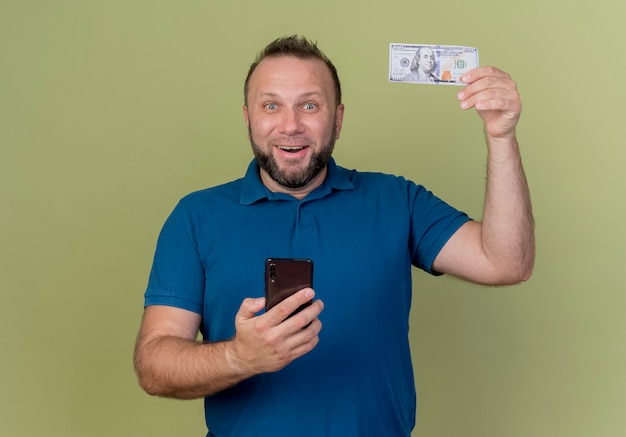 Joyful adult slavic man raising money and holding mobile phone 