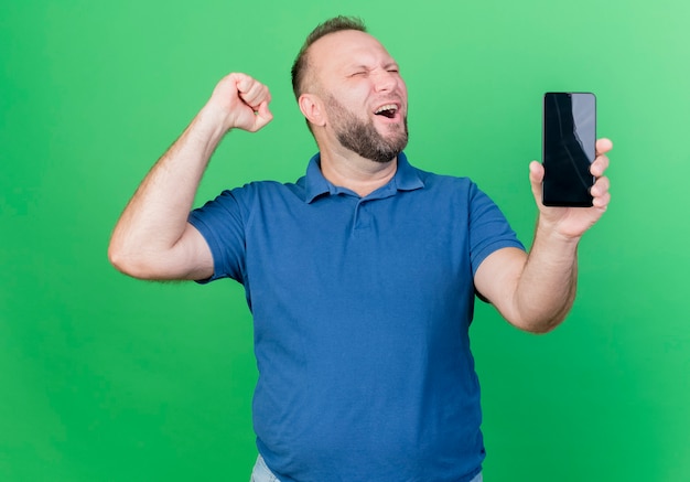 Joyful adult slavic man showing mobile phone and doing yes gesture with closed eyes isolated on green wall