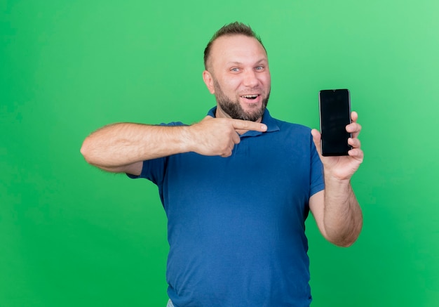 Радостный взрослый славянский мужчина показывает и указывает на мобильный телефон, изолированный на зеленой стене