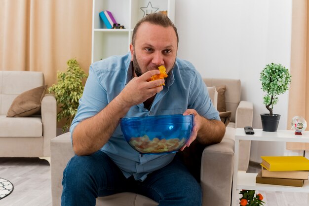 Joyful adult slavic man sits on armchair holding and eating bowl of chips inside the living room
