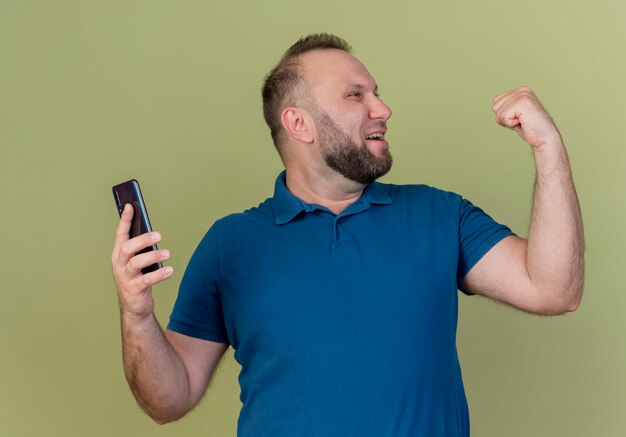 Joyful adult slavic man turning head to side holding mobile phone doing yes gesture 