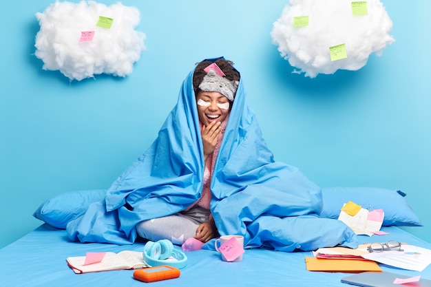 無料写真 うれしそうなアフリカ系アメリカ人の女の子が幸せに笑うベッドの上で柔らかい毛布のポーズに包まれた明るい気分が試験の準備をしています