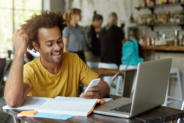 Радостный афро-американский студент сидит за деревянным столом в кафе в окружении книг, тетрадей, ноутбука, держа в руке сотовый телефон и смотрит с удовольствием