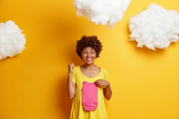 Joyful African American woman clenches fist and feels happy as finds out she will have daughter, holds baby singlet over tummy, stands against yellow  with clouds above. Pregnancy concept
