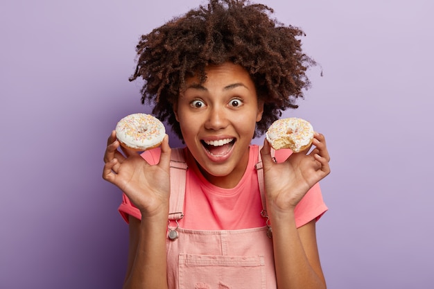Joyful African American woman has delicious pastry breakfast, holds two sweet glazed doughnuts, enjoys tasty dessert, eats unhealthy food, isolated against purple wall. Female sweet tooth