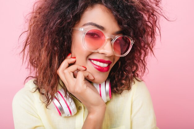 Joyful african female model with red lips smiling, touching her face. Close-up portrait of lovable curly lady in sunglasses and headphones laughing with pleasure.