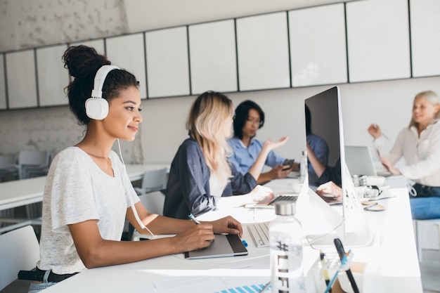 Joyful african web-developer working on new project while listening music in white headphones. Black female designer doing her job in office with talking colleagues.