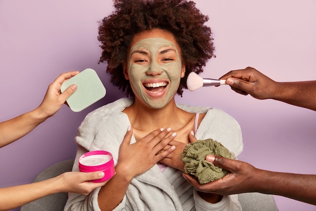 Joyful Afro American woman laughs sincerely, applies peeling mask, gets different beauty treatments at same time