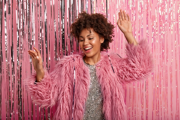 joyful Afro American woman laughs sincerely, feels relaxed, dances while listens favorite music, wears pink fur coat and sparkling dress, models over rosy wall. Celebration