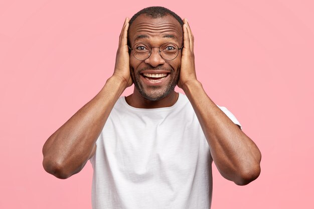 Joyful amazed man recieves congratulations, keeps both hands on head, has broad friendly smile, isolated over pink wall