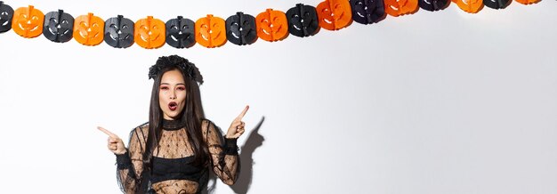 Joyful asian woman in gothic lace dress celebrating halloween pointing fingers sideways standing aga