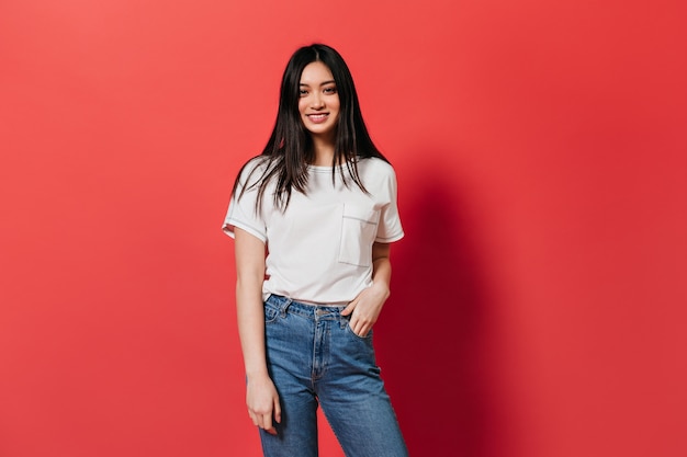 Joyful Asian woman in jeans and t-shirt looks into front on isolated wall