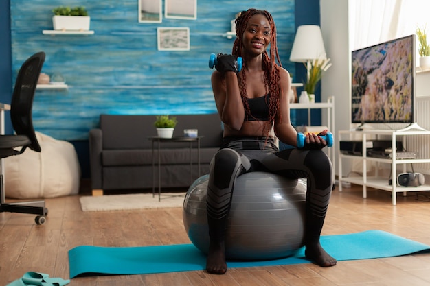 Joyful athletic black person in leggings using stability ball for training biceps, doing curls with dumbbells. Strong athletic person doing sports at home using modern equipment for