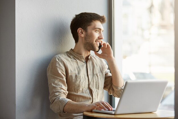 즐거운 매력적인 면도 남자 coworking 공간에 앉아, 랩톱 컴퓨터에서 작업 하 고 저녁에 날짜에 대 한 여자 친구와 전화 통화.