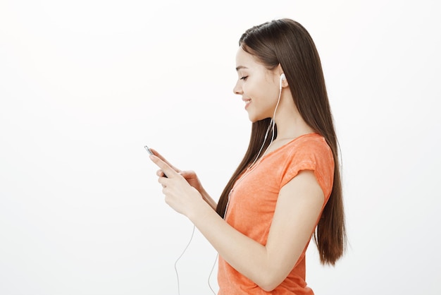 Радостная привлекательная женщина, стоящая в профиле на сером фоне, держит смартфон, глядя на экран с чувственной заботливой улыбкой, слушая музыку в наушниках
