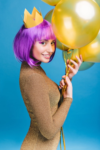 Joyful attractive young woman with cut purple hair having fun with golden balloons . Crown on head, makeup with tinsels, luxury fashionable dress, celebrating new year party.