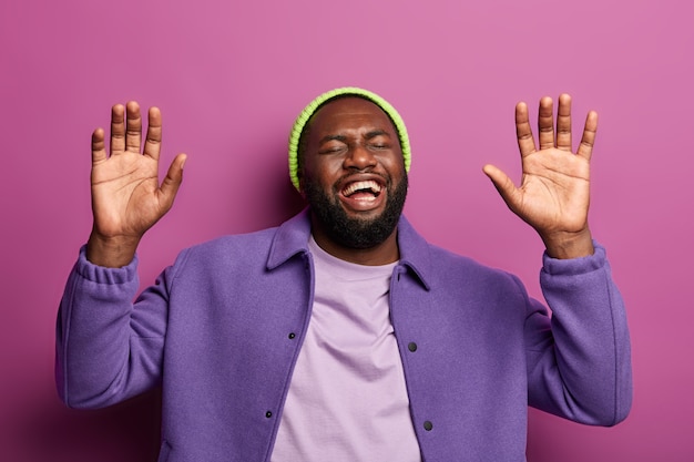 Free photo joyful bearded afro american man raises palms, cannot stop laughing, hears funny anecdote, feels overemotive, watches hilarious comedy or joke