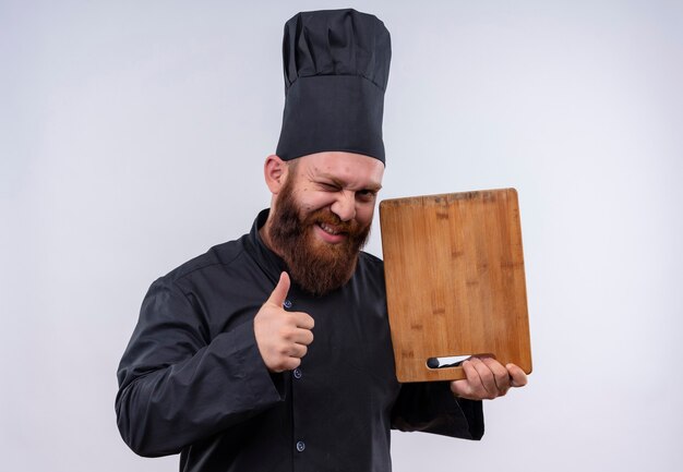 A joyful bearded chef man in black uniform showing wooden kitchen board with thumbs up on a white wall