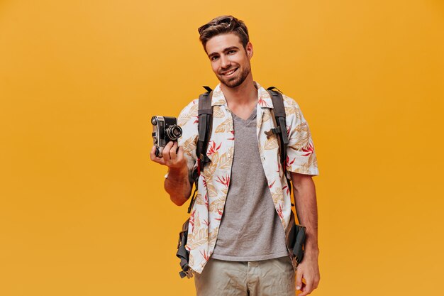 Joyful bearded guy in grey t-shirt and fashionable printed shirt smiling and holding camera on isolated orange wall